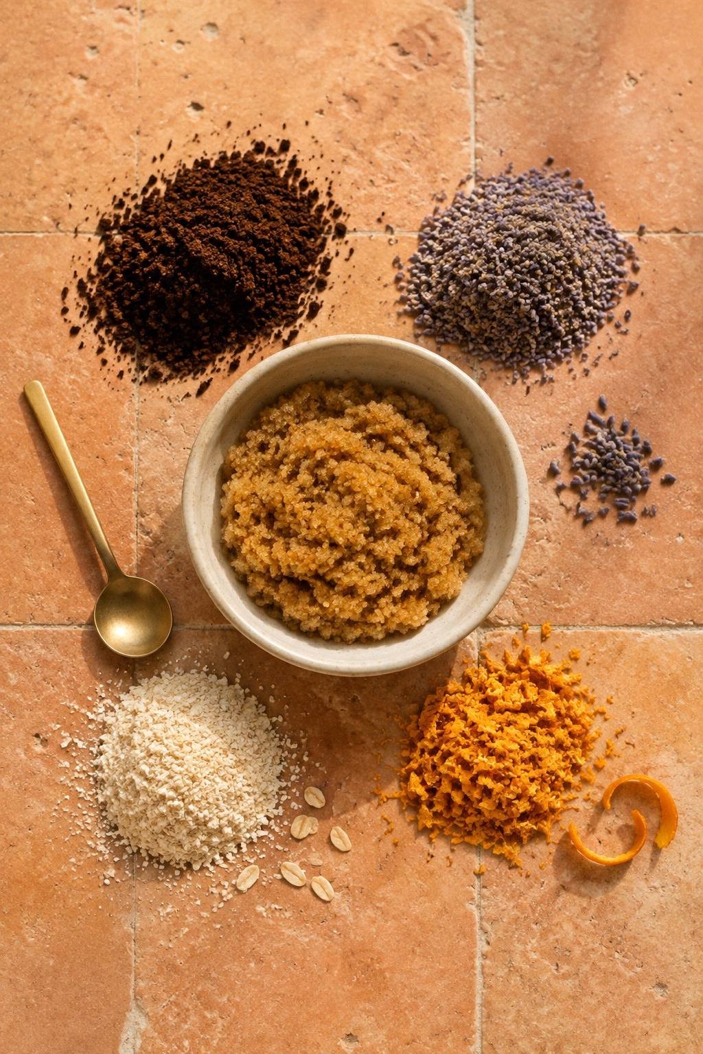 Dried coffee grounds, ground dried lavender, ground oats, and dried orange zest arranged around a bowl of plain sugar scrub on terracotta tile
