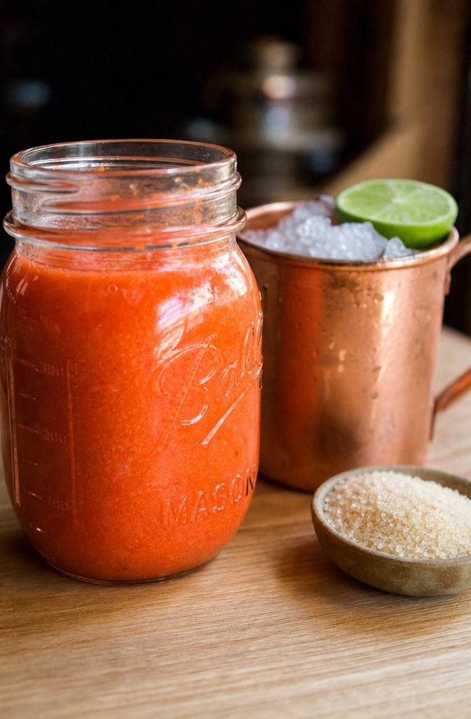 Glass jar of bright orange peach puree on a light wood counter beside a halved lime and a copper mug filled with crushed ice ready for a peach moscow mule mocktail